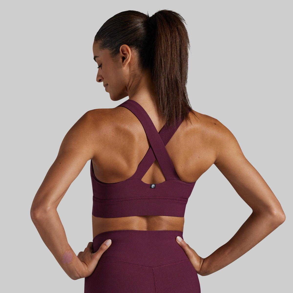 A woman shown from the back wearing a ribbed athletic sports bra and matching high-waisted leggings in a deep plum purple color, set against a solid light gray background.