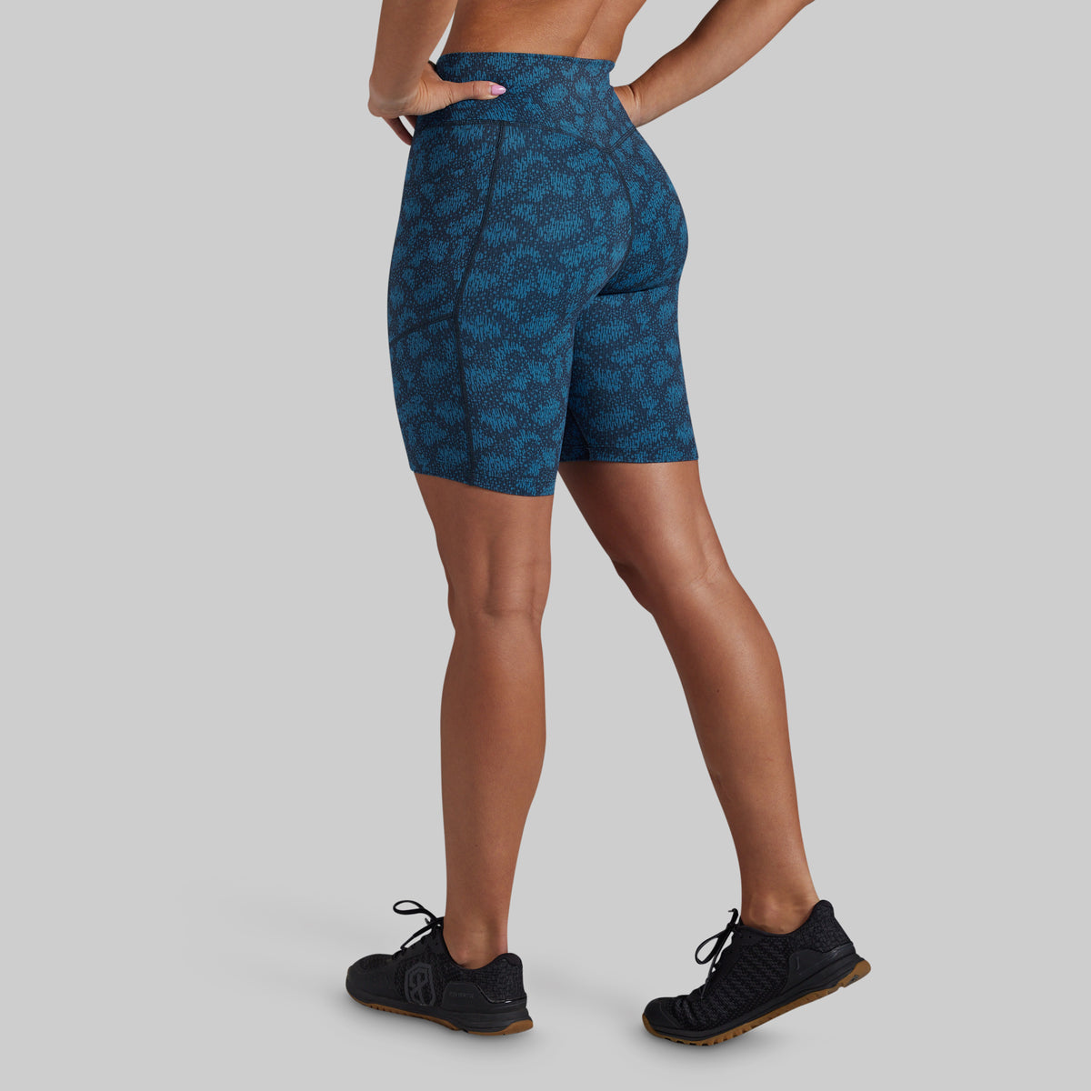 A model from the back wearing high-waisted athletic biker shorts with a dark blue and teal abstract pattern, paired with black athletic shoes, against a solid light gray background.
