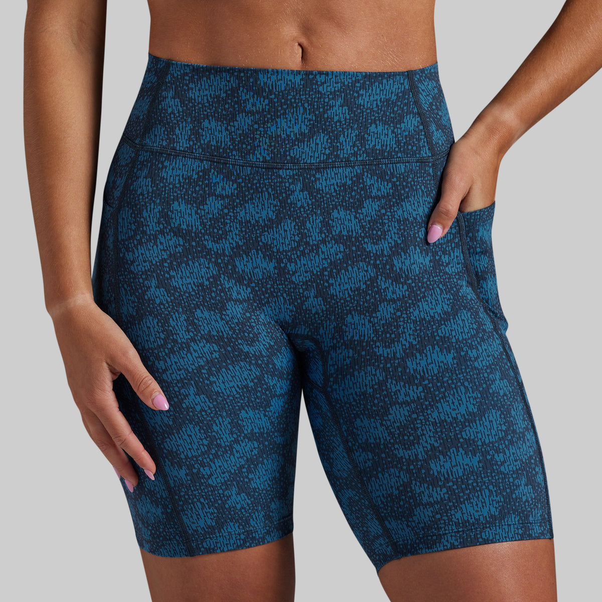 A close-up of a model wearing high-waisted athletic biker shorts with a blue and black abstract pattern, shown from the waist to mid-thigh against a solid gray background.