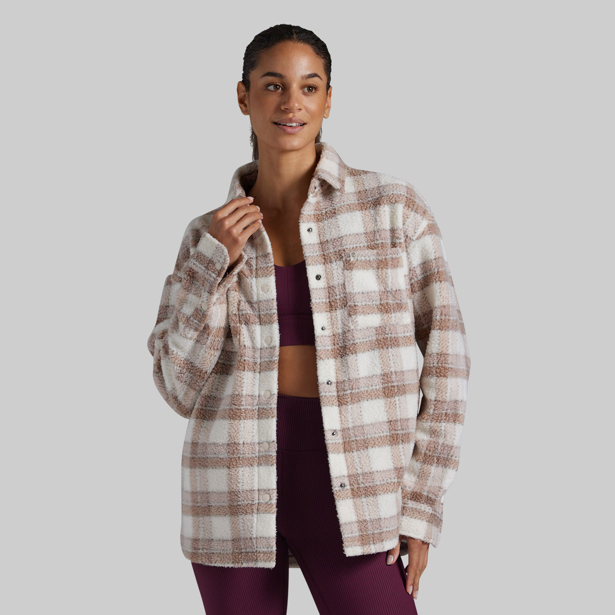 A woman modeling a long-sleeve button-front plaid overshirt in cream and taupe, worn open over a dark plum crop top and matching leggings, against a solid light gray studio background.
