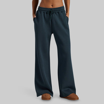 A person wearing dark teal wide-leg sweatpants with an elastic drawstring waistband and side pockets, paired with brown slippers, shown against a plain light gray studio background.