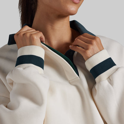 Close-up of a person wearing a cream-colored knit quarter-zip sweatshirt with dark teal ribbed trim on the collar and cuffs, shown against a light gray studio background.