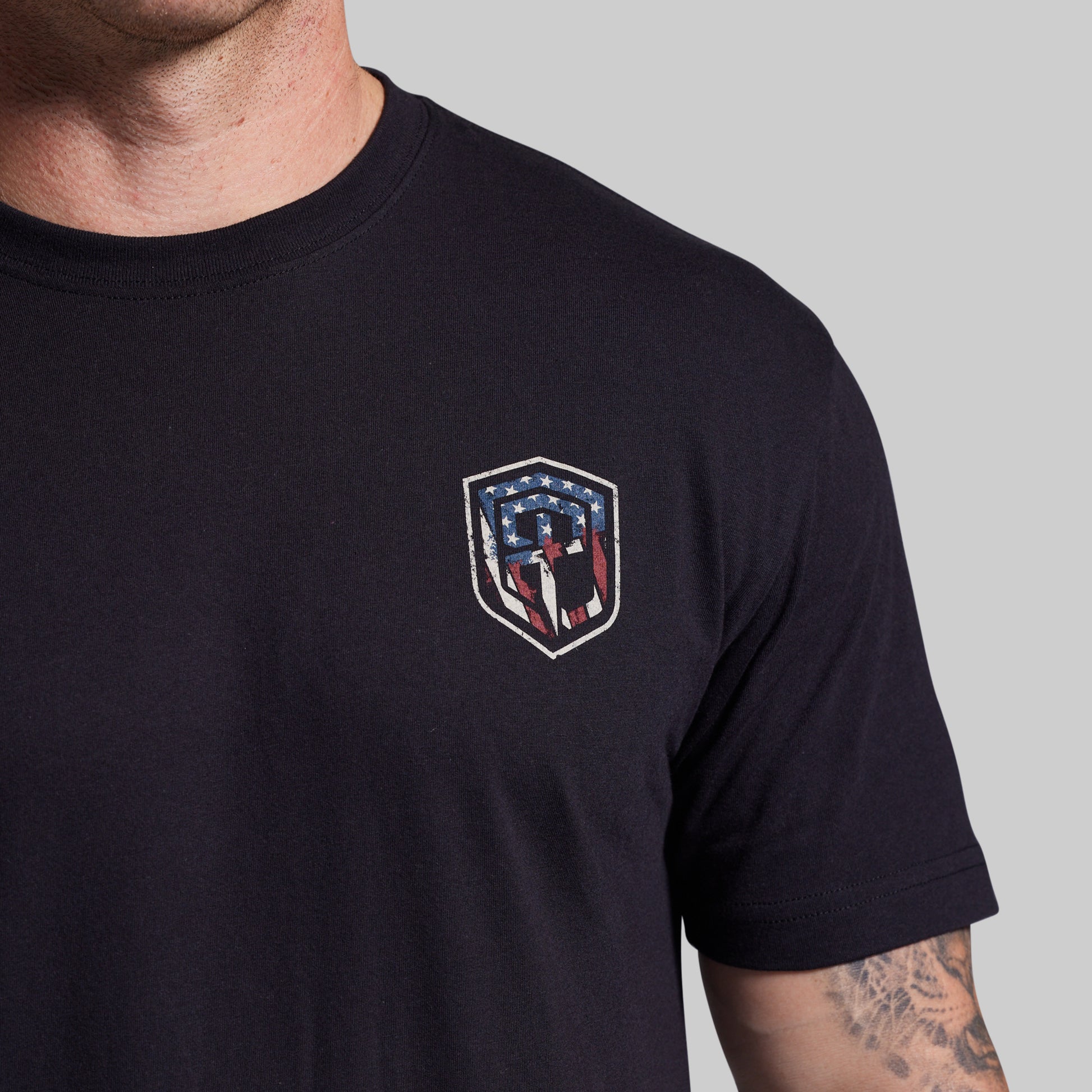 Black t-shirt with a shield emblem featuring American flag colors on a gray background