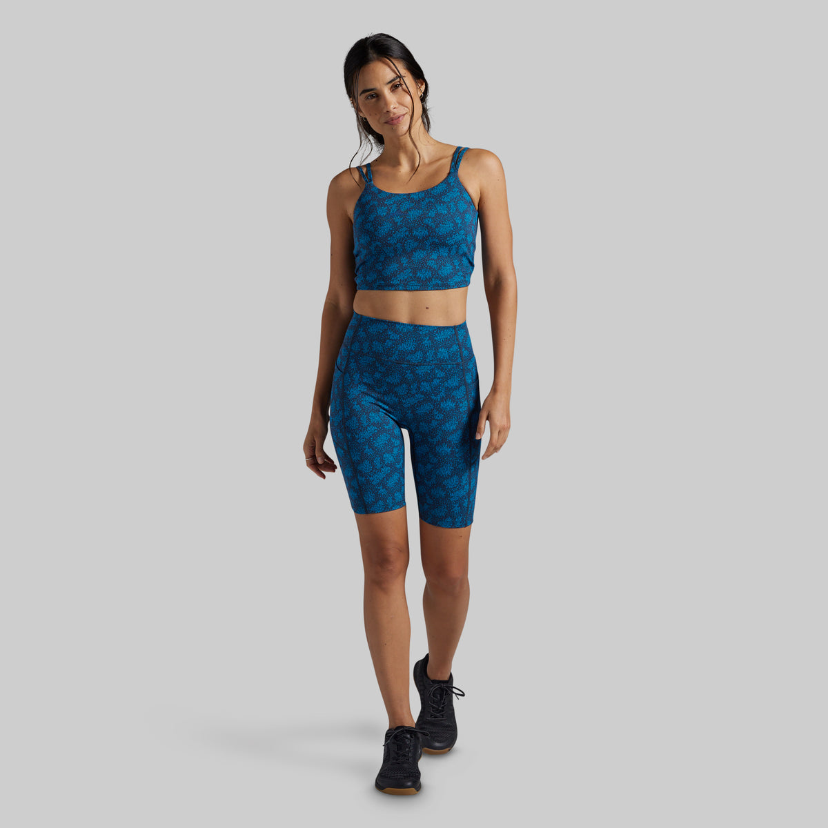 A woman modeling a matching athletic set consisting of a blue patterned sports bra and high-waisted biker shorts, paired with black sneakers, against a solid gray background.