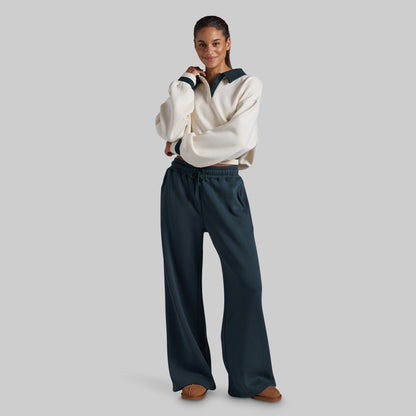 A woman modeling a cream sweatshirt with dark teal trim and matching dark teal wide-leg sweatpants, standing against a solid light gray studio background.