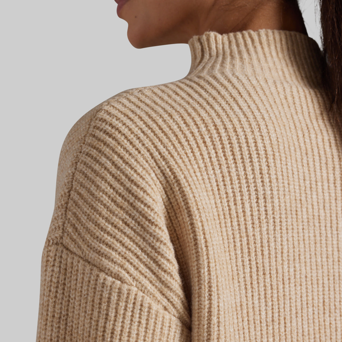 Close-up of a person wearing a ribbed knit mock neck sweater in a warm beige color, showing the texture and shoulder detail against a light gray background.