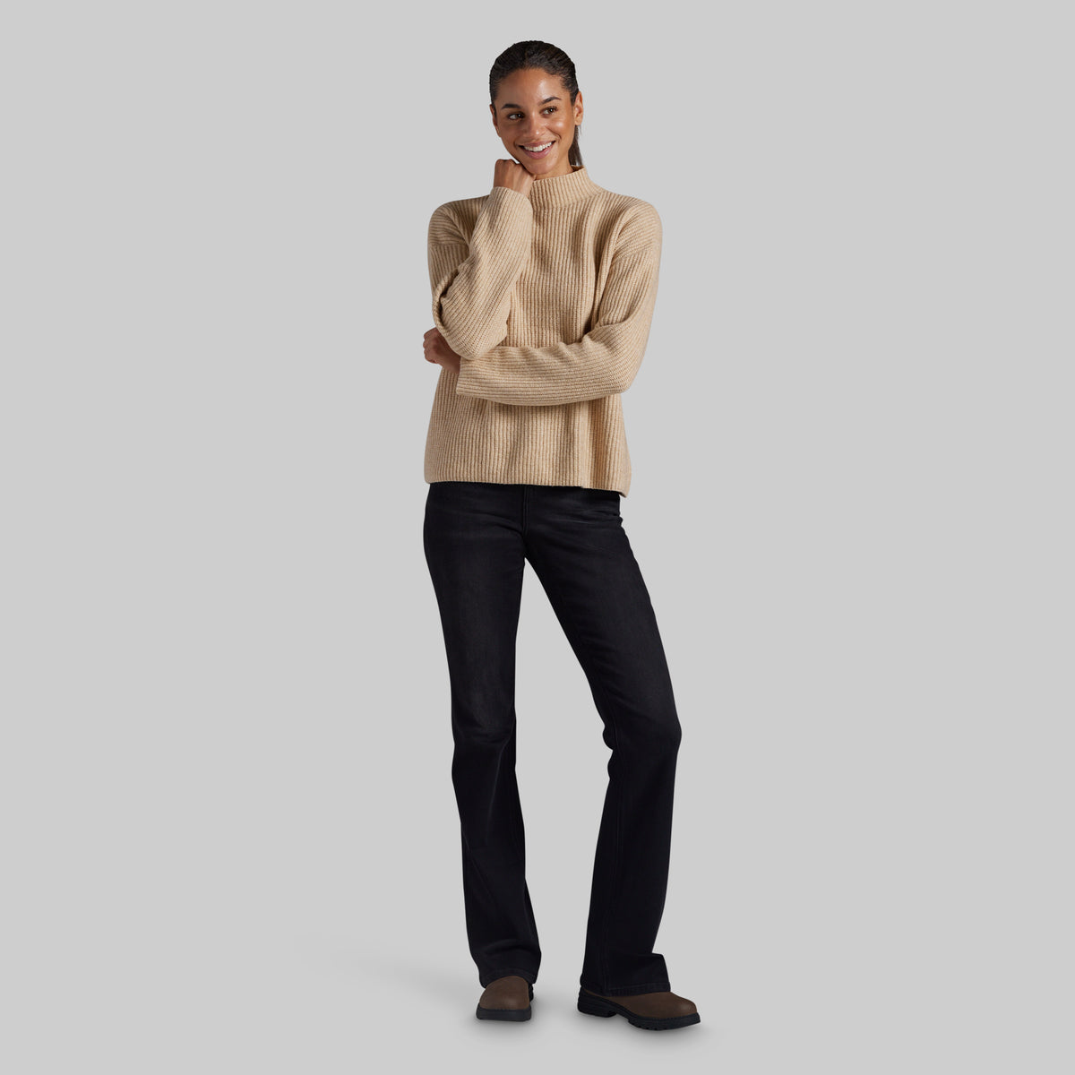 A woman modeling a beige ribbed knit mock-neck sweater and black jeans against a solid light gray background.
