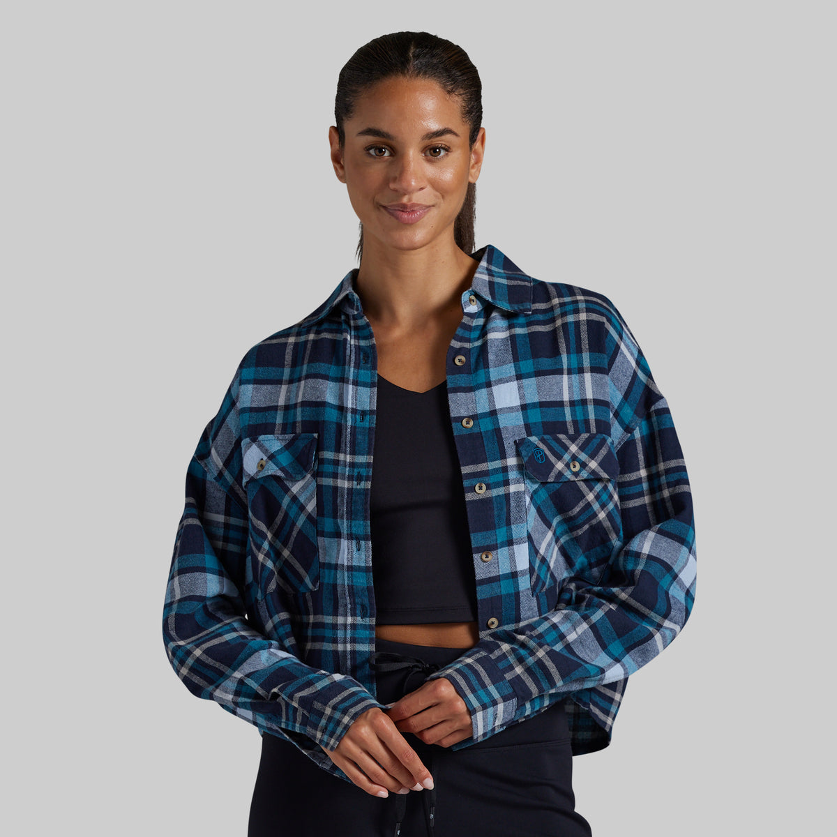 A woman modeling a cropped, long-sleeve plaid button-up shirt in shades of navy, blue, and white over a black crop top, set against a solid gray studio background.