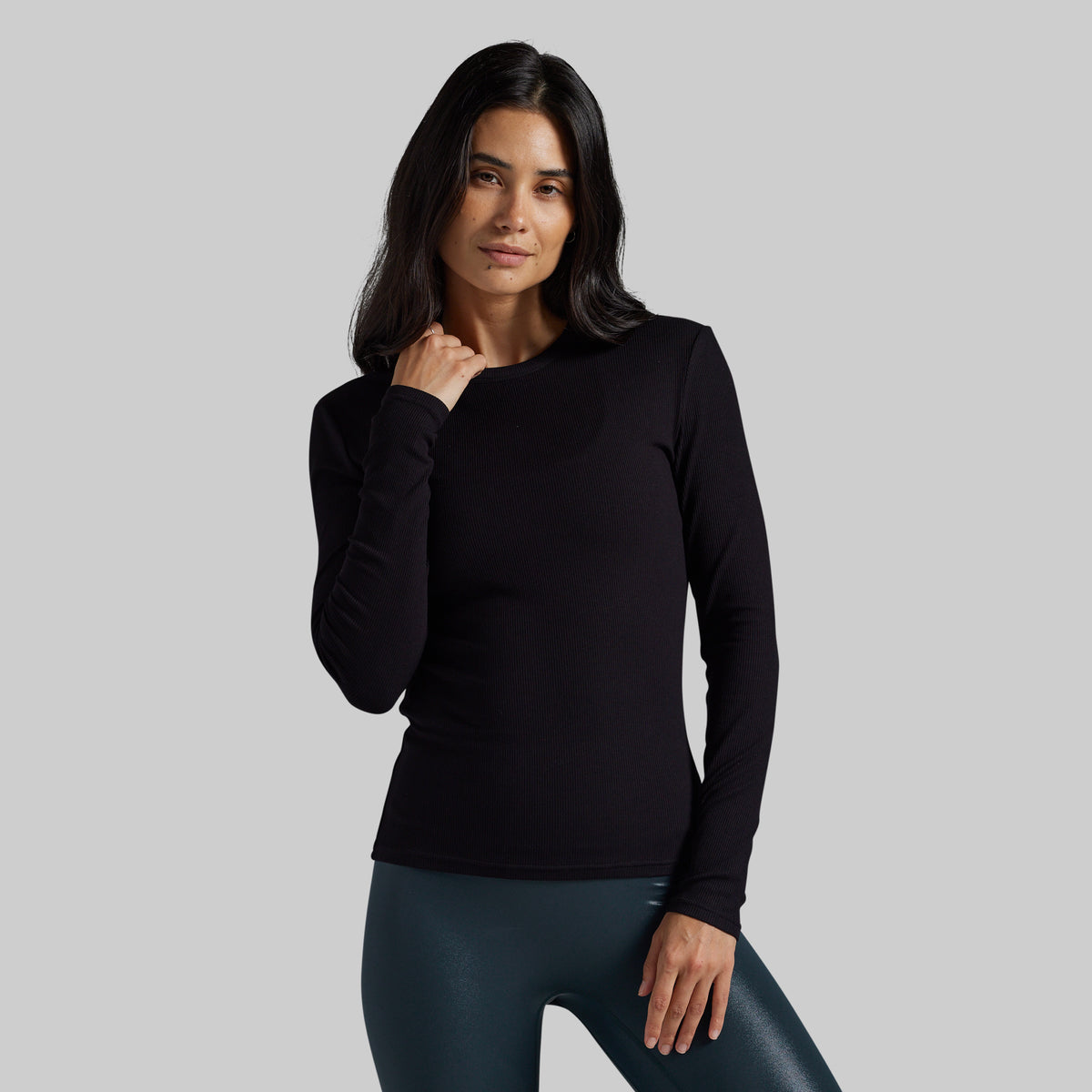 A woman modeling a black long-sleeve crewneck top against a solid gray background.
