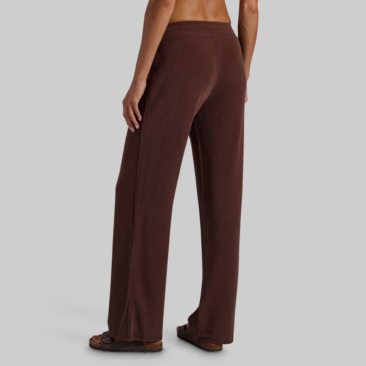 A person shown from the back wearing loose, wide-leg brown pants and brown sandals against a plain light gray studio background.