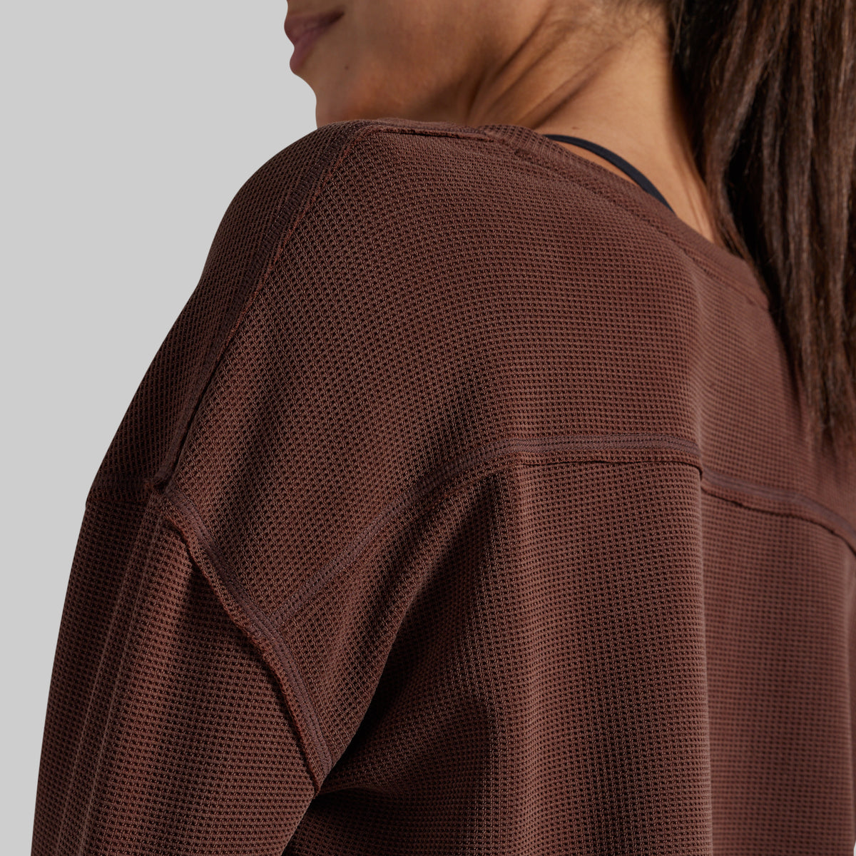 Close-up of a woman wearing a brown textured long-sleeve top, showing the shoulder, neck, and hair against a solid gray background.