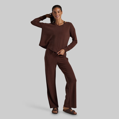 A woman modeling a matching dark brown lounge set consisting of a long-sleeve cropped top and wide-leg pants, paired with brown sandals, against a solid light gray background.