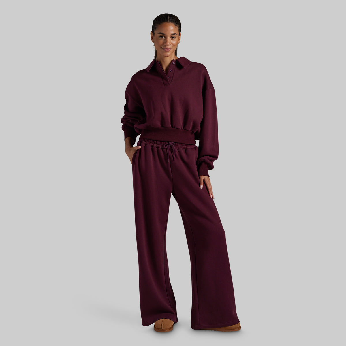 A model wearing wide-leg lounge pants in deep burgundy with a matching collared sweatshirt, paired with brown suede slippers, against a solid gray studio background.