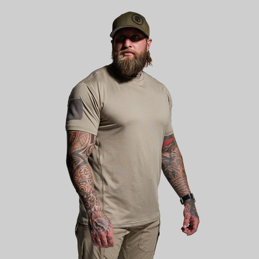 Man wearing a beige t-shirt and cap with tattoos on arms against a gray background