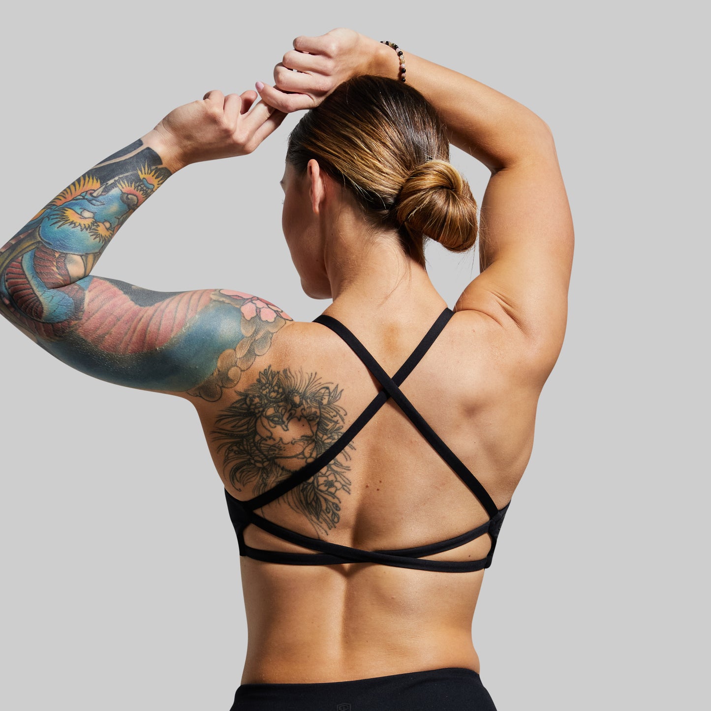 Person wearing a black strappy sports bra with tattoos on arms and back, against a plain background