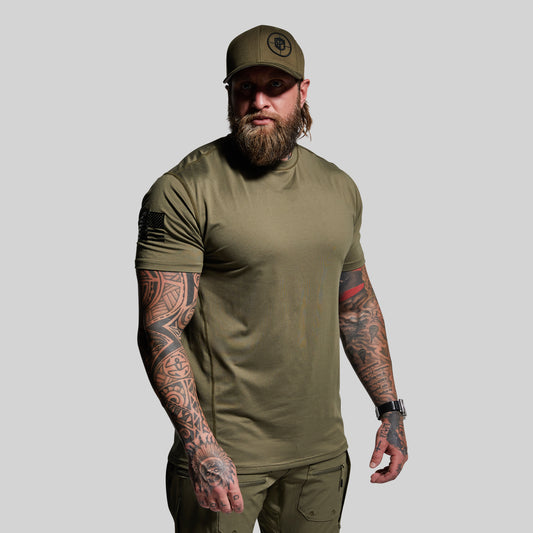 Man wearing an olive green t-shirt, cap, and pants with tattoos on arms and hands against a gray background