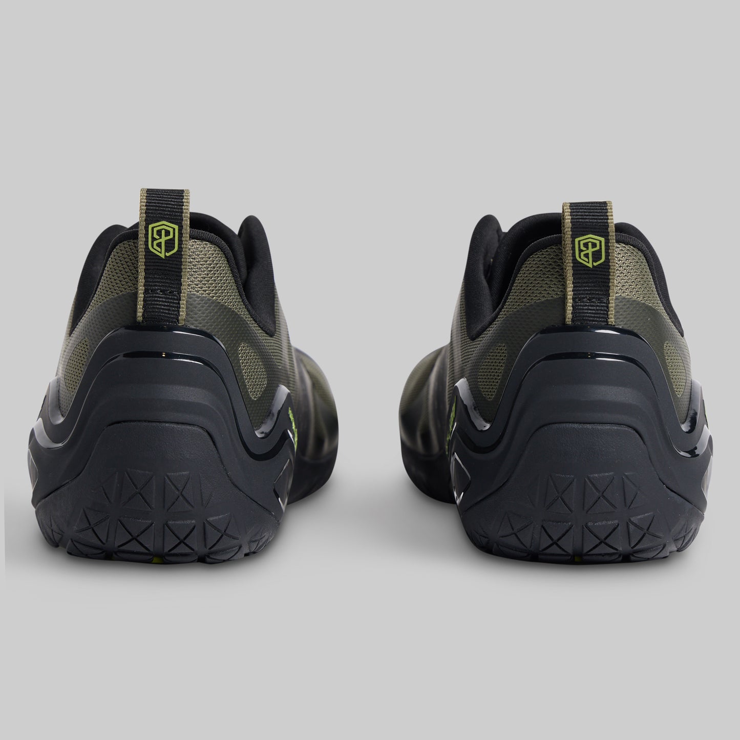 A pair of athletic sneakers shown from the rear, featuring a black and olive green design with a small yellow-green shield logo on the heel pull tabs, set against a solid gray background.