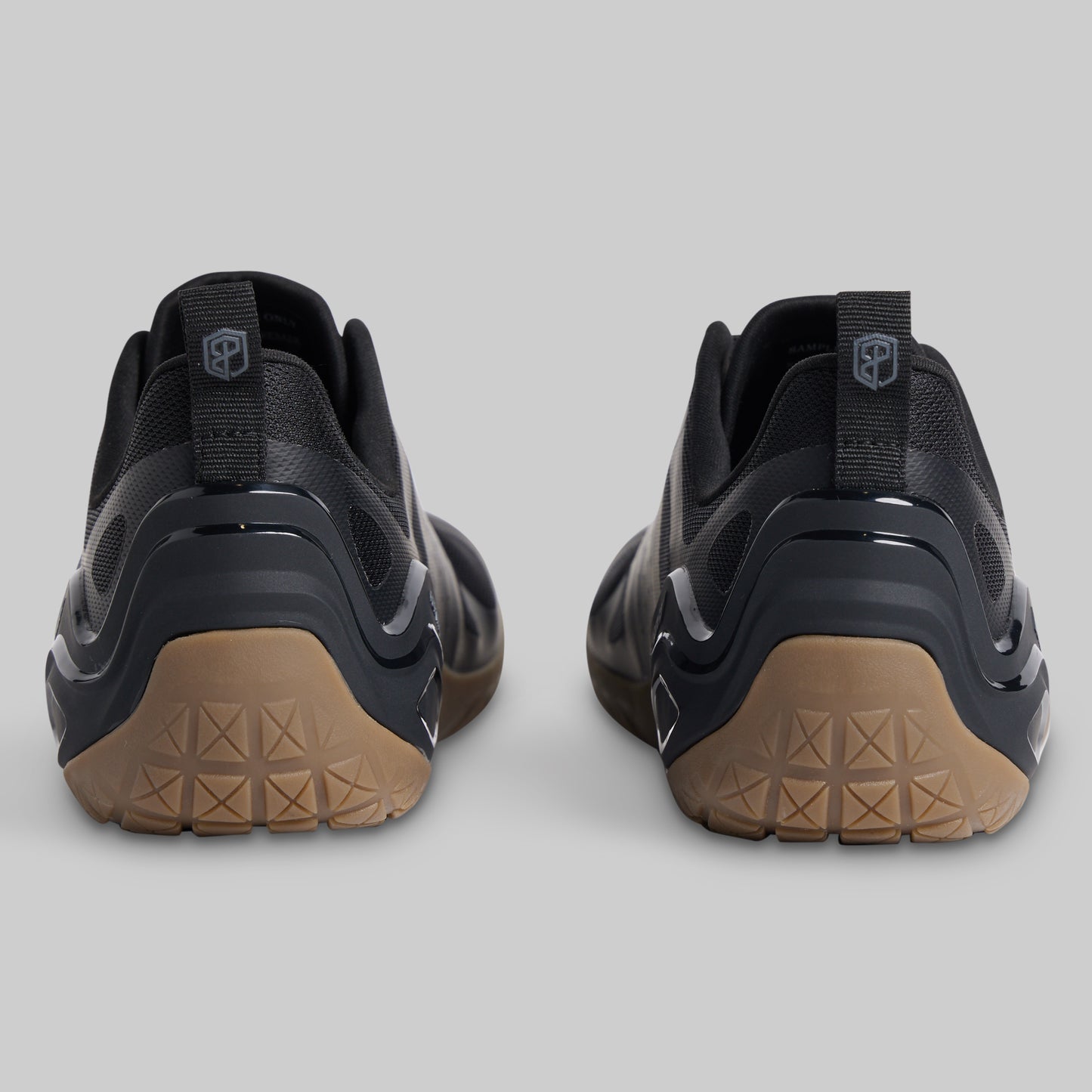 A pair of black minimalist sneakers shown from the rear, featuring textured tan outsoles and a pull tab with a shield logo on a light gray background.