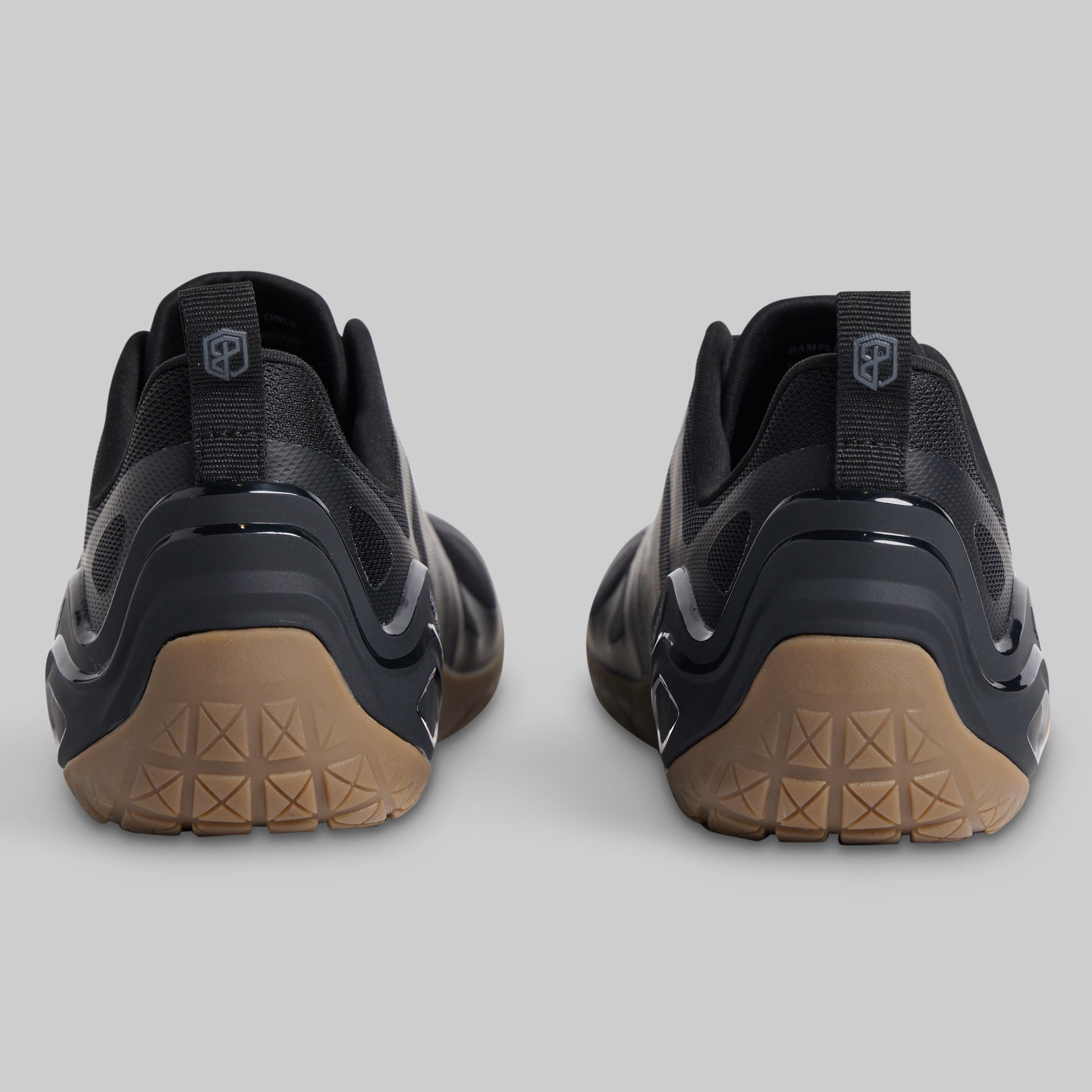 A pair of black minimalist sneakers shown from the rear, featuring textured tan outsoles and a pull tab with a shield logo on a light gray background.