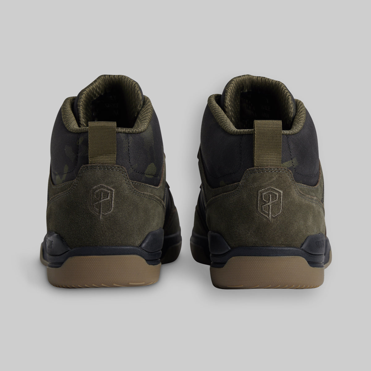 A pair of mid-top sneakers shown from the back, featuring olive green suede and black camo panels with a tan sole, set against a solid light gray background.