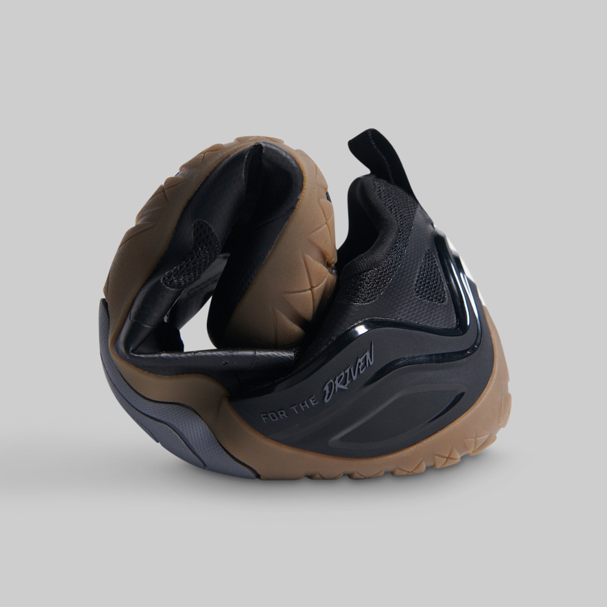 A black and brown athletic barefoot shoe rolled to show flexibility against a solid gray background.