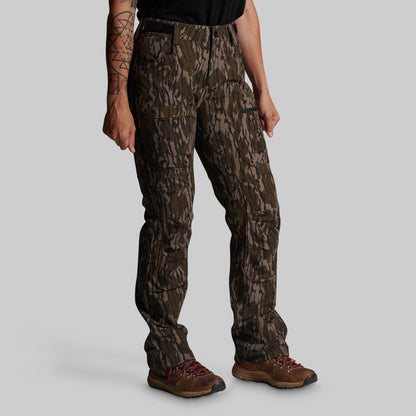 A person wearing camouflage cargo pants with a brown shirt and brown hiking boots, shown from the chest down against a plain light gray studio background.