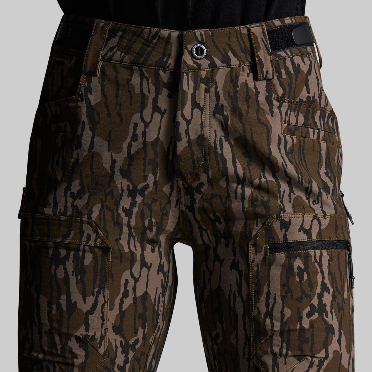Close-up of a pair of cargo pants featuring a brown and black mossy oak camouflage pattern, shown on a model wearing a black top against a solid gray background.