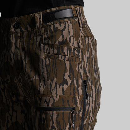 Close-up of a pair of camouflage outdoor pants worn by a model, showing the side pocket, zipper, belt loop, and waistband against a solid gray background.