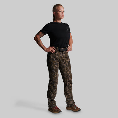 A woman modeling a pair of brown and black camouflage hunting pants, paired with a black t-shirt, black belt, and brown hiking boots, against a solid gray background.