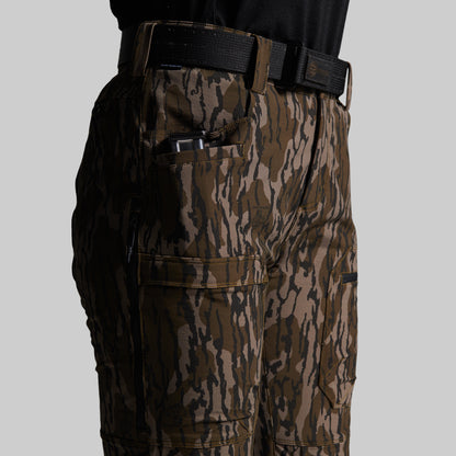 Close-up of a person wearing outdoor cargo pants with a brown and black bark camouflage pattern, paired with a black belt and black shirt, against a solid gray background.