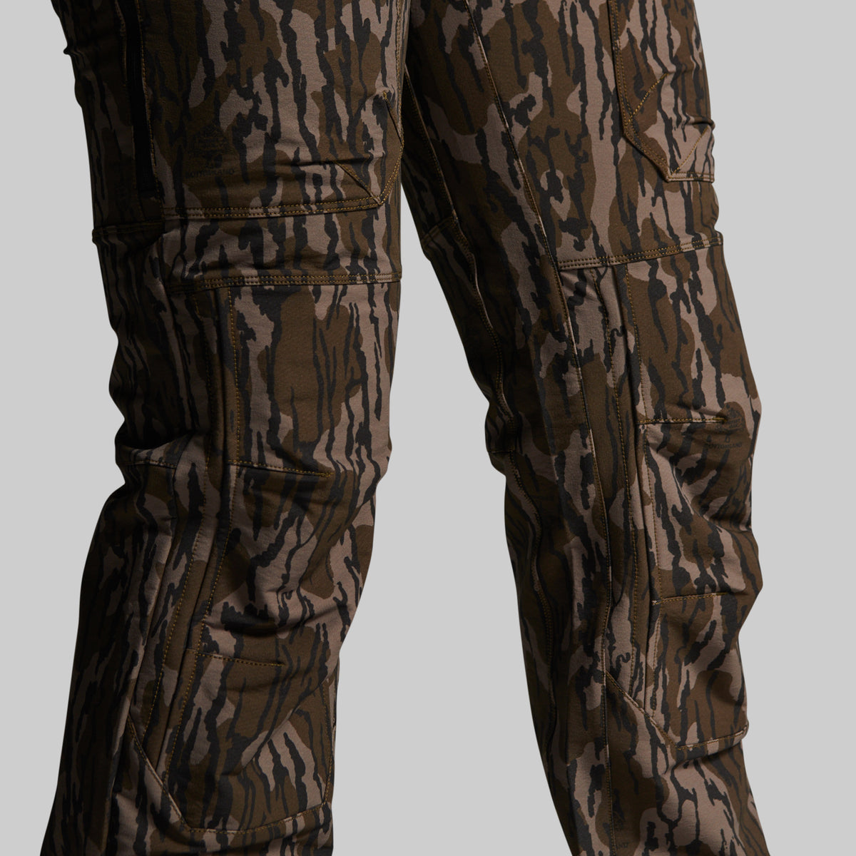 Close-up of camouflage cargo pants on a model, showing the lower torso and legs with multiple zippered and flap pockets against a solid gray background.