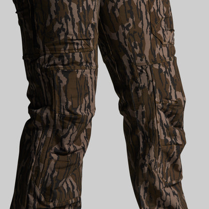 Close-up of camouflage cargo pants on a model, showing the lower torso and legs with multiple zippered and flap pockets against a solid gray background.