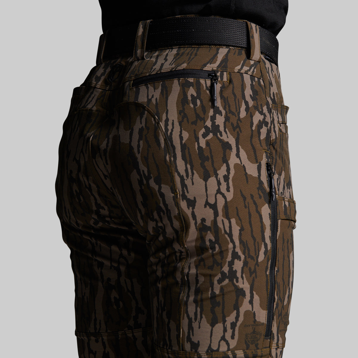 Close-up of the back and side of camouflage outdoor pants with a black belt, shown against a light gray studio background.