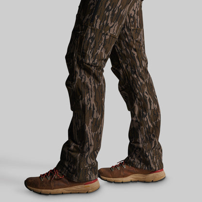 Close-up of a person's lower legs wearing brown camouflage pants and brown hiking boots against a solid gray background.