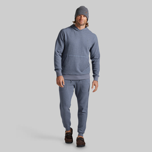 A man modeling a matching heathered slate blue hoodie and jogger sweatpants set, paired with a gray beanie and brown shoes, against a solid gray background.