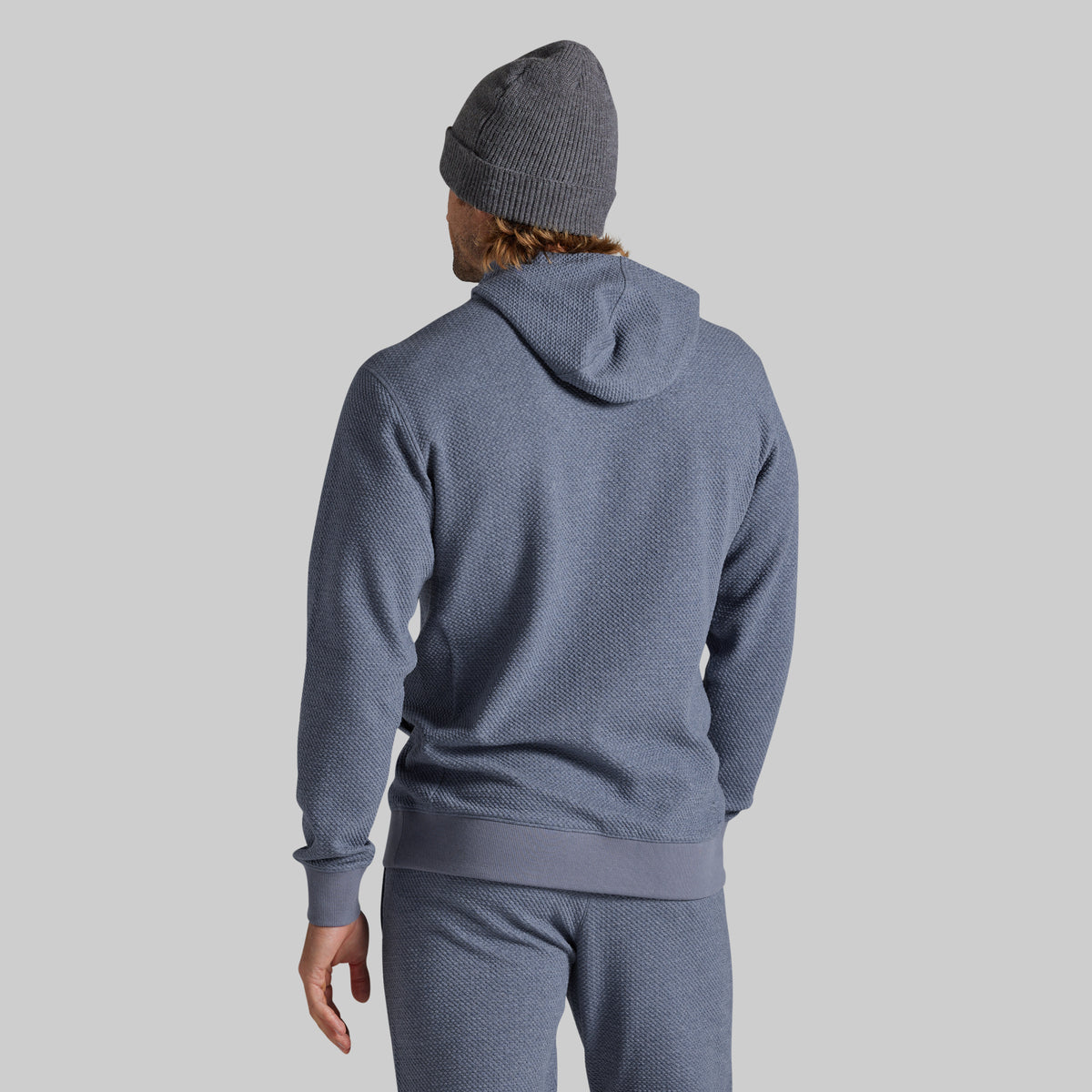 A man wearing a heathered slate blue hoodie and matching sweatpants, viewed from behind, wearing a gray knit beanie against a solid light gray background.