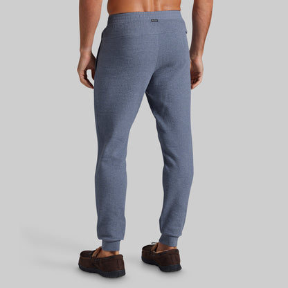 A man viewed from the back wearing textured blue jogger pants and brown suede loafers against a solid gray studio background.