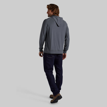 A man is shown from behind wearing a slate gray hooded sweatshirt, dark navy pants, and brown shoes, standing against a solid light gray background.