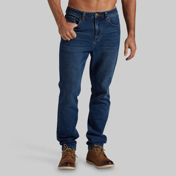 A man wearing dark blue slim-fit jeans and brown leather boots, shown from the waist down against a solid light gray background.