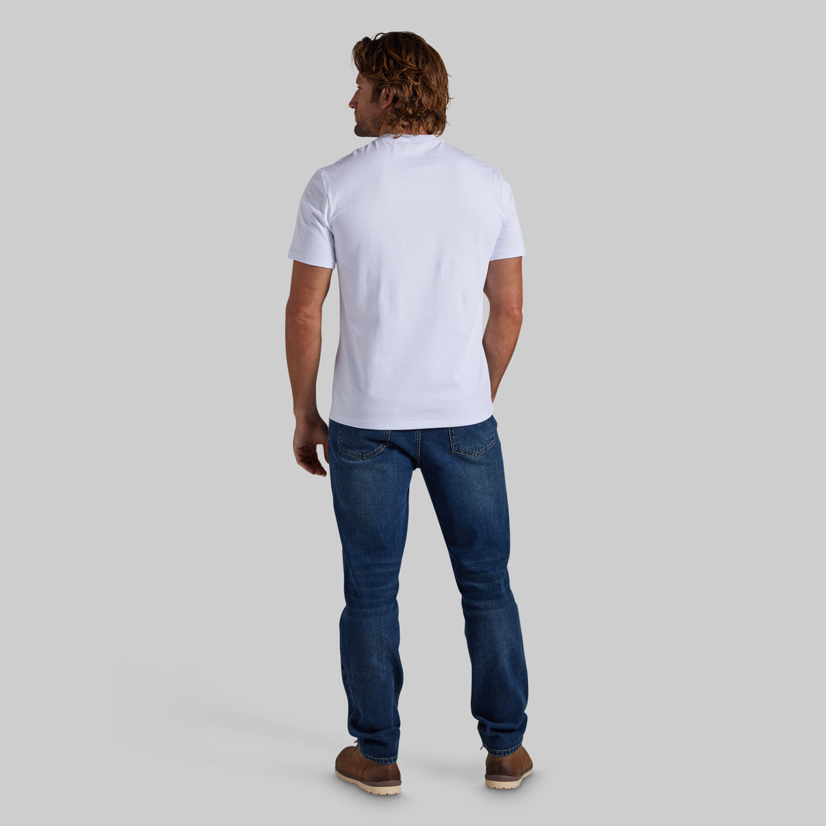 A man viewed from behind wearing a white short-sleeve shirt, dark blue jeans, and brown shoes, standing against a solid light gray background.