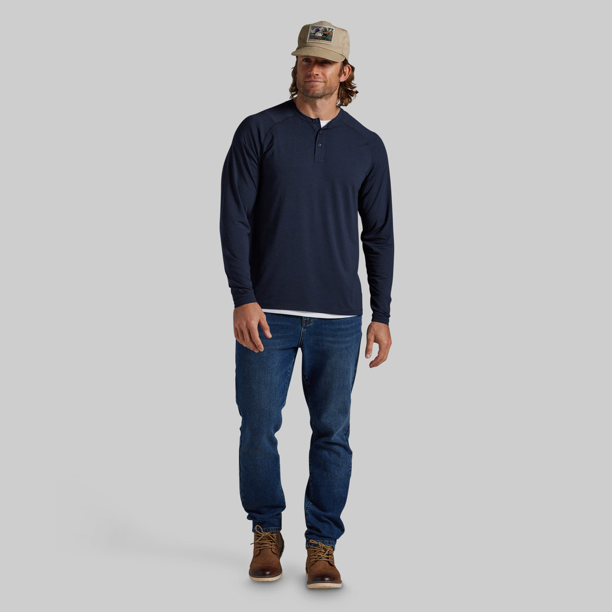 A man modeling a navy blue long-sleeve henley shirt, blue jeans, brown boots, and a tan baseball cap against a solid gray background.