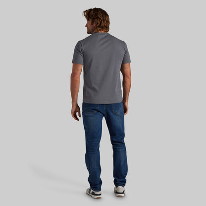 A man viewed from behind wearing a dark gray short-sleeve t-shirt, blue jeans, and white sneakers, standing against a solid gray studio background.