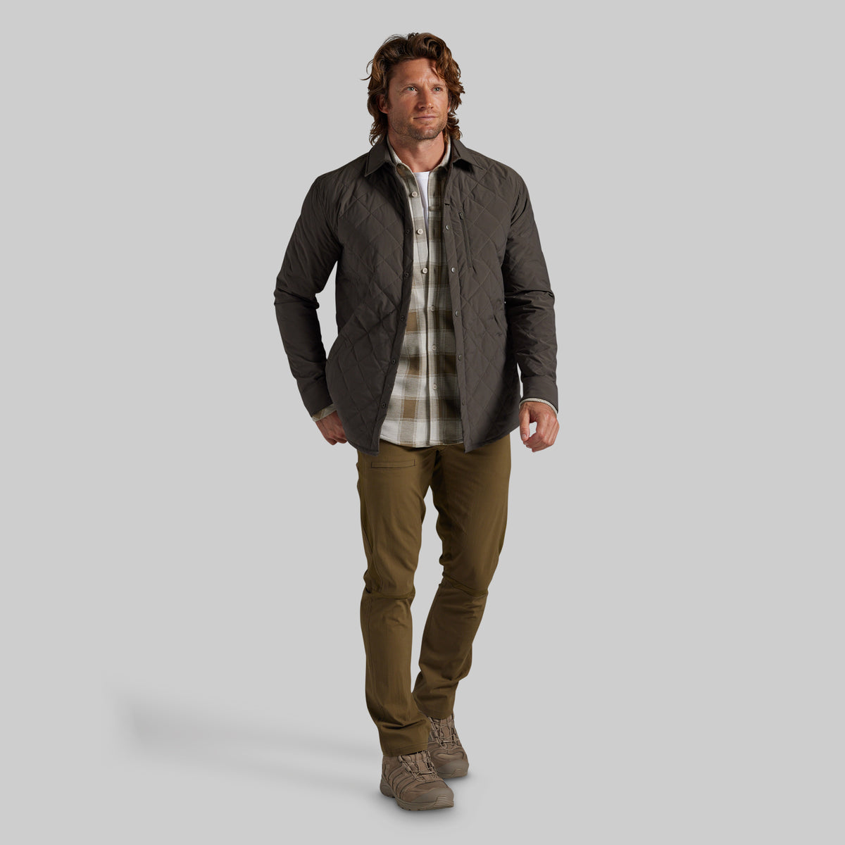 A man modeling a dark gray quilted jacket over a beige and brown plaid flannel shirt, paired with olive green pants and brown boots, against a solid gray background.