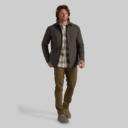 A man modeling a dark gray quilted jacket over a beige and brown plaid flannel shirt, paired with olive green pants and brown boots, against a solid gray background.