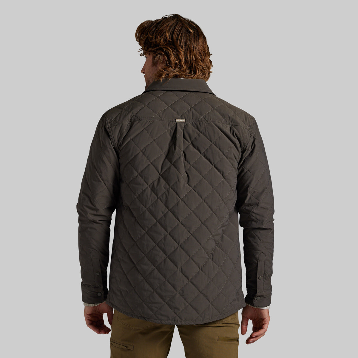 A man viewed from the back wearing a dark gray quilted button-up shirt jacket, paired with olive green pants, against a solid gray studio background.
