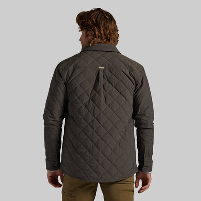 A man viewed from the back wearing a dark gray quilted button-up shirt jacket, paired with olive green pants, against a solid gray studio background.