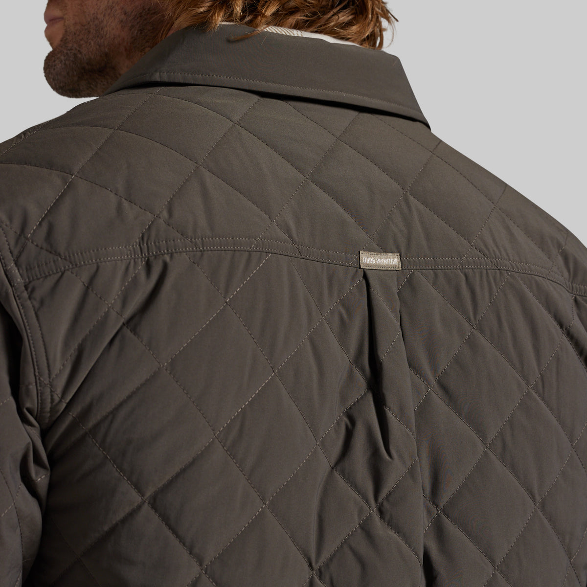 Close-up back view of a man wearing a dark olive quilted jacket, showing the diamond stitching and a small rectangular brand tag on the upper back against a light gray background.