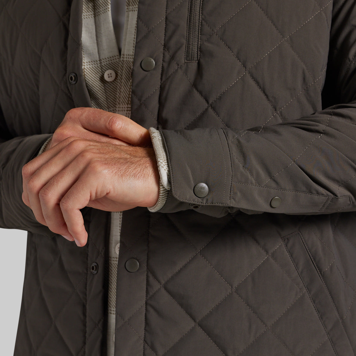 Close-up of a person wearing a dark grey diamond-quilted jacket, showing the quilted texture, snap-button closure, and a plaid shirt underneath.