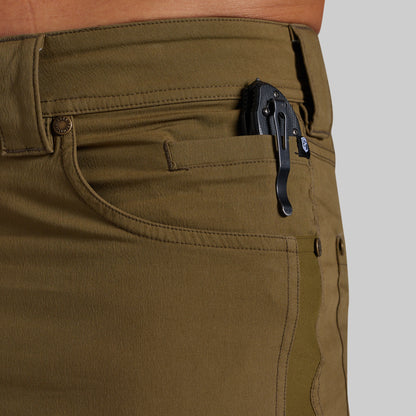 Close-up of olive green pants showing the back pocket with a black clip holding a black knife, worn by a model against a light gray background.