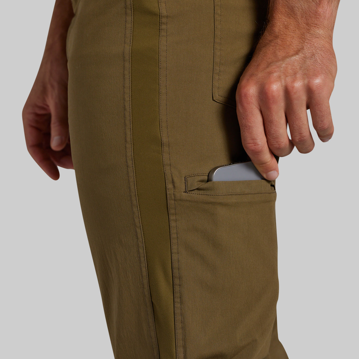 Close-up of a person wearing olive green pants, showing a side pocket with a gray smartphone partially tucked inside, against a light gray background.